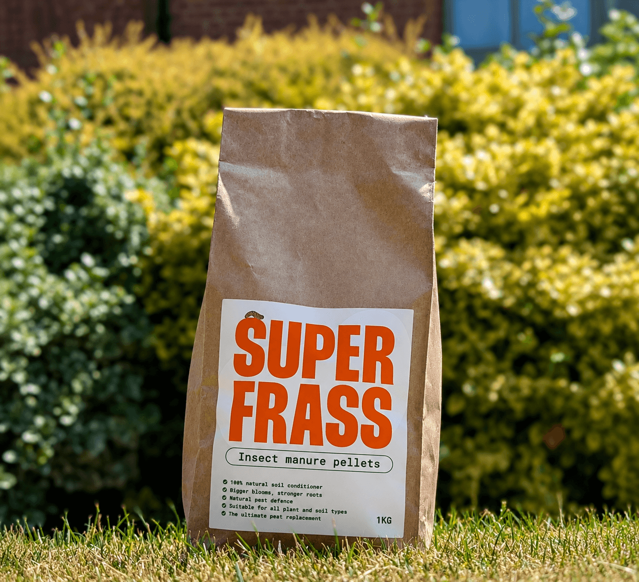 Superfrass insect manure pellets 1kg bag standing on a lawn with garden planting behind it